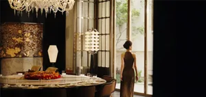 A woman stands in front of a window in a large, elegant dining room with a wooden dining table and chairs, surrounded by wine glasses