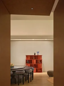 A modern study with a wooden floor, a dining table, a red bookshelf, and a chair, lit by natural sunlight