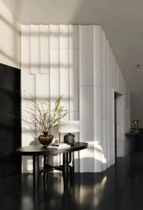 A modern dining area with a black table and a vase, surrounded by a large white wall and a doorway, with natural light streaming in