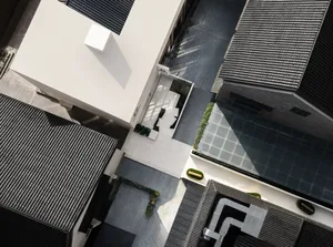 A modern building with a white concrete facade, a balcony, and a patio, surrounded by a courtyard, with natural sunlight and a bright atmosphere