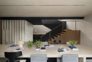 A well-lit living room with a dining table, chairs, and a staircase, featuring a cozy and inviting atmosphere with books and magazines scattered on the table