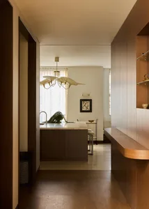 A modern kitchen with wooden floors, cabinets, and a dining table, lit by natural light from the windows, with a potted plant and chairs in the space