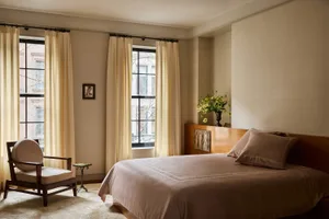 Cozy bedroom with a bed, chair, and window, featuring a pink blanket, flowers, and natural light