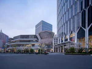 城市街道景觀，背景為大型建築，兩旁林木排列，停放汽車、行人及自行車