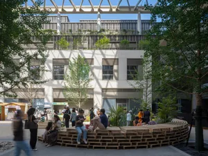 A bustling public space with a wooden bench in the center, surrounded by a modern building with a mix of concrete and wood, and people interacting in the outdoor area