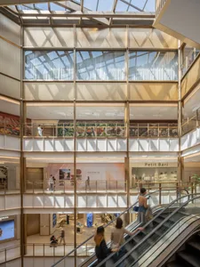 A large, open commercial space with a glass ceiling, natural light, and potted plants, filled with people walking around