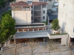 A modern building with a large glass wall and rooftop terrace, featuring a furnished outdoor seating area with chairs and dining tables, set against a concrete facade in a natural daylight setting