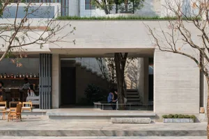 A modern house with a concrete exterior, a patio area with a dining table and chairs, and a tree in the front yard, set in a minimalist and contemporary style