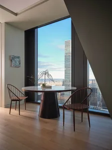 Cozy modern living space with a dining table, chairs, and a potted plant, surrounded by a large window with a city view, and well-lit with natural light