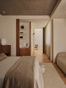 A calm and inviting bedroom with a bed, chair, and couch, decorated with wooden furniture and a white color scheme, with natural light streaming in through the window
