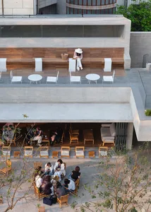 A casual outdoor restaurant with wooden furniture, a woman sitting on a bench, and other people in the scene
