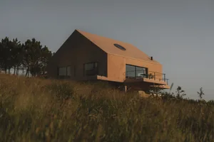 A modern house on a hill with a large window, surrounded by natural scenery and bathed in warm sunlight