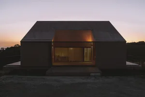 A modern house with a brown exterior, a large window, and a grassy area, photographed at dusk, creating a warm and inviting atmosphere