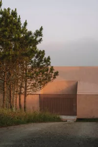 A modern building with a pink exterior, surrounded by trees, featuring a large door and window, with a concrete floor and a bench in a serene outdoor setting
