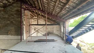 A man kneeling on a dirt floor in a construction site with a metal beam and cement floor, focused on his task