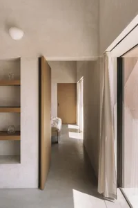 A cozy bedroom with a wooden floor, a white wall, a bed, a bookshelf, and a vase on the bookshelf, with natural light entering through an open door