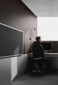 A man sits at a desk in a small office space with a wooden floor and a window, working on a computer