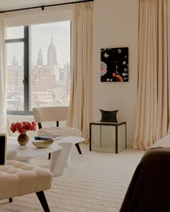 A modern living room with a large window overlooking the city, featuring a couch, chair, and dining table, with a vase and potted plant adding greenery to the space, lit by natural light
