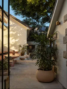A residential patio with a dining table and chairs, surrounded by a potted plant, with a house in the background, featuring a mix of wood and stone elements, and natural sunlight creating a warm atmosphere