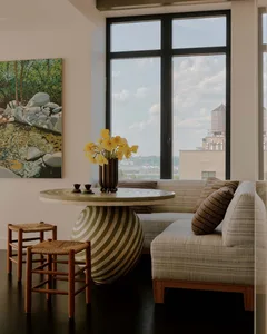 A well-lit living room with a couch, chair, and dining table, featuring a vase with yellow flowers and a large window for natural light