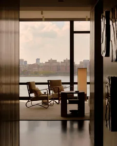 A modern living room with a couch and chair near a large window overlooking a city, a dining table on the right, and a bright and inviting atmosphere