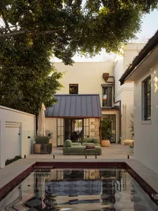 A house with a white exterior and a backyard featuring a pool with a blue tiled bottom, surrounded by a patio with a couch and chair, and a dining table and potted plant