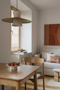A modern living room with a wooden dining table, chairs, and couch, featuring a bowl of fruit and natural light from a window