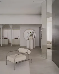 A modern room with a white color scheme, a circular window, and natural light, featuring a couch, chair, bench, and potted plants