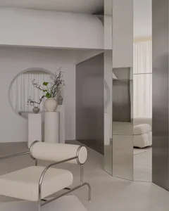 A modern living room with a large mirror, a couch, a chair, and a potted plant, featuring white and grey tones and soft inviting lighting