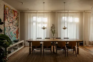 A modern dining room with a wooden table, chairs, and a vase with flowers, surrounded by natural light and a warm atmosphere
