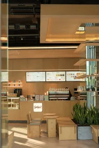 A modern restaurant interior with a dining area and kitchen, featuring wooden elements, a coffee machine, and a refrigerator, with soft and warm lighting
