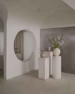 A minimalist living space with white walls, a large mirror, and a vase with flowers on a pedestal, set against a white floor