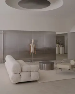 A modern living room with a white couch, chair, and mannequin, surrounded by white walls and silver-toned accents, with natural light pouring in through the windows
