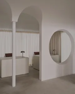 A modern bathroom with two sinks on a counter, a chair, and a round mirror, set against a predominantly white background with dim lighting creating a cozy atmosphere
