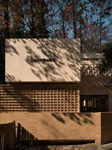 Photograph of a modern building with a tan exterior, featuring a large brick wall with a window and a door, set in a natural environment with trees and warm sunlight