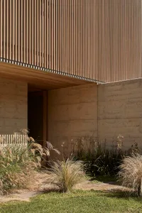 A modern house with a concrete exterior, large window, and door, surrounded by a garden with green plants, bathed in natural light