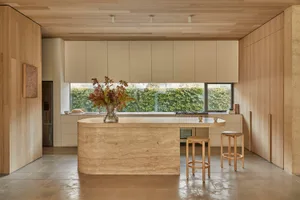 A well-lit kitchen with a marble countertop, wooden cabinets, a sink, refrigerator, and stools, creating a bright and inviting atmosphere