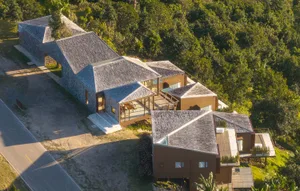 A modern house with a triangular roof, surrounded by trees, featuring a well-lit interior with a dining table and chairs, and a blend of natural materials and indoor-outdoor spaces