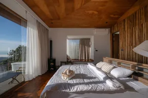 A simple bedroom with a bed, chair, and window, featuring a wooden floor and white walls, well-lit by natural light