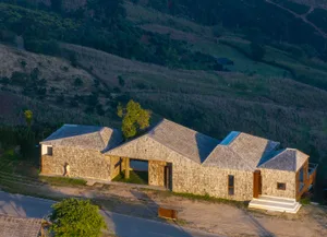 A stone house with a triangular design and large window sits on a hillside in a rural area surrounded by a lush green field