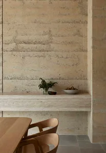 A modern dining area with a wooden table and chairs, surrounded by a stone wall and countertop, with a cozy dim lighting and a potted plant nearby
