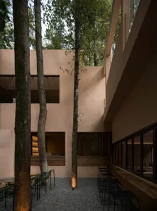 A modern building with a large patio area surrounded by trees, featuring a concrete exterior, a dining table, and chairs, with natural light and a relaxing atmosphere