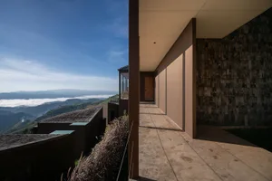 A modern house with a brown exterior, situated on a hill, featuring a large window and door, with natural light and a serene atmosphere