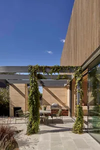 Outdoor dining space with a table, chairs, potted plants, and a vase, surrounded by a rustic stone and wood setting with a fireplace and natural sunlight