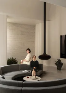 Two women sitting on a couch in a well-lit modern living room with a dining table and potted plant, surrounded by white materials