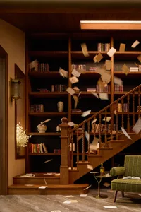 A cluttered room with a staircase, wooden railing, and a bookshelf with scattered books, creating a sense of disarray