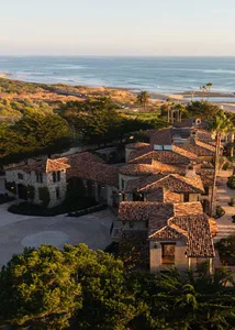 A large Spanish-style house with a red tile roof and various windows, situated on a hillside overlooking the ocean, bathed in natural light