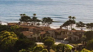 A large Spanish-style house with a red roof and white stucco walls, surrounded by lush greenery and overlooking the ocean