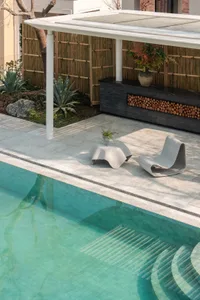 A serene outdoor patio area with a pool, wooden fence, and potted plants, featuring a bench and chair for relaxation