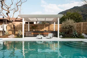 A serene pool area with a pool house, surrounded by a fence and potted plants, featuring a bench, chair, and dining table